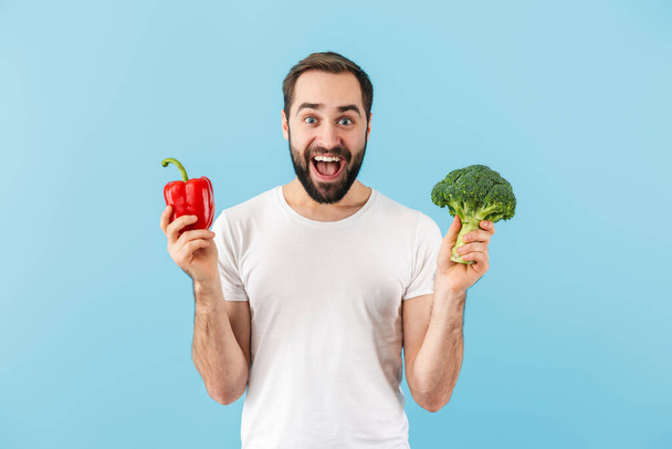 Handsome cheerful young bearded man wearing t-shirt standing isolated over blue background, showing red capsicum - Photo, image