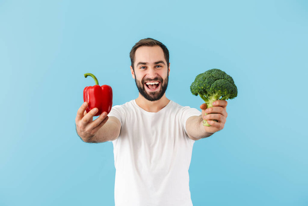Handsome cheerful young bearded man wearing t-shirt standing isolated over blue background, showing red capsicum - Φωτογραφία, εικόνα