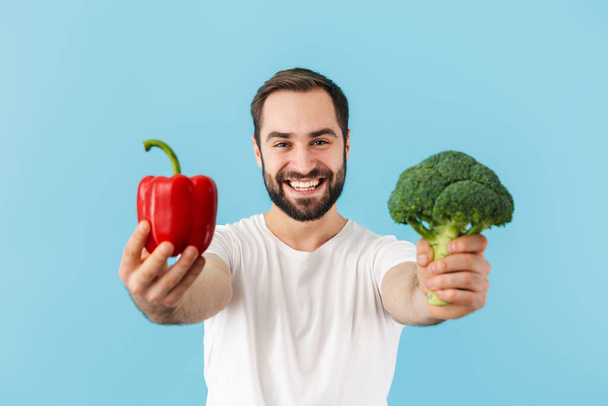 Handsome cheerful young bearded man wearing t-shirt standing isolated over blue background, showing red capsicum - Foto, afbeelding
