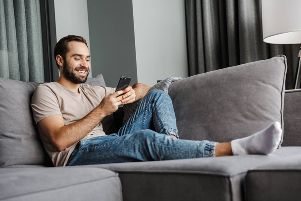 Image of a handsome cheerful happy young positive man indoors at home on sofa using mobile phone. - Foto, immagini