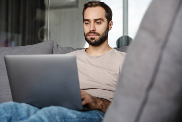 Image of a concentrated young man indoors at home on sofa using laptop computer. - Foto, imagen