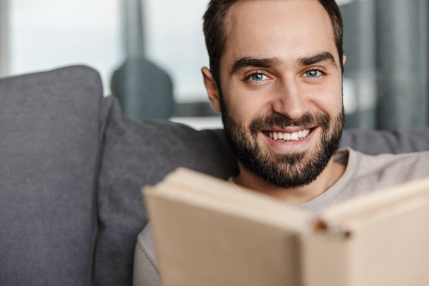 Image of a happy positive young man indoors at home on sofa reading book. - Foto, imagen