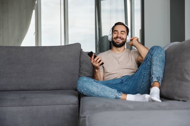 Image of a happy young positive man indoors at home on sofa listening music with headphones using mobile phone. - Foto, afbeelding