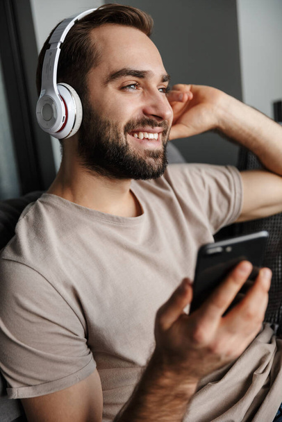 Afbeelding van een glimlachende optimistische jonge knappe man binnen op de bank luistermuziek met koptelefoon met mobiele telefoon. - Foto, afbeelding