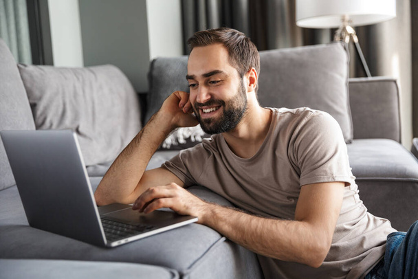 Image of a cheerful optimistic young man indoors at home on sofa using laptop computer. - Foto, immagini