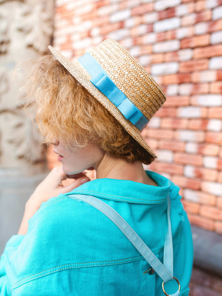 νέα χαρούμενη κατσαρομάλλα κοκκινομάλλα με μπλε τζιν μπουφάν και ψάθινο καπέλο ταξιδιώτη. Διασκέδαση, καλοκαίρι, ταξίδια, μόδα, έννοια της νεολαίας - Φωτογραφία, εικόνα
