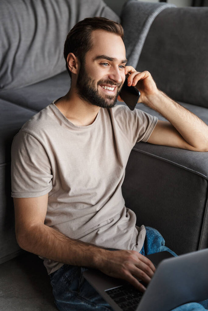 Image of a cheery pleased young man indoors at home on sofa using laptop computer talking by mobile phone. - Photo, Image