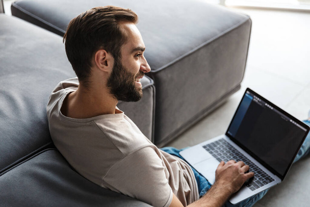 Image of a cheerful optimistic young man indoors at home on sofa using laptop computer. - Foto, imagen