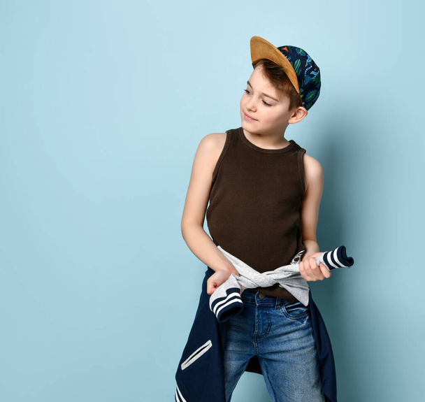 Teenage boy in cap, brown undershirt and jeans. He is tying hoodie around his waist and smiling. Posing on blue studio background - Photo, image