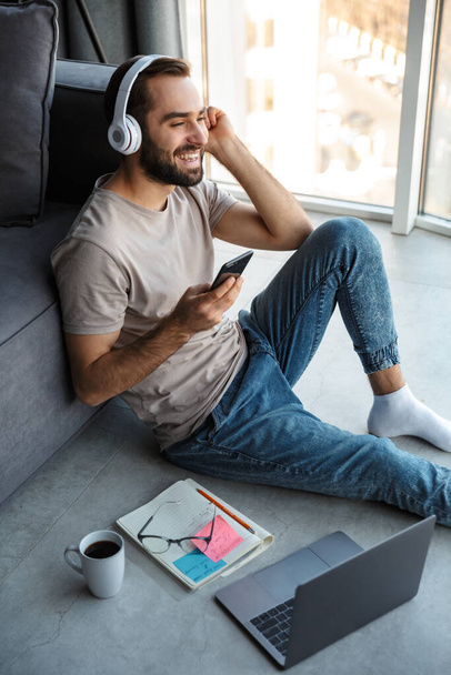 Attractive smart young man sitting on a floor in the living room, listening to music with wireless headphones and mobile phone - Фото, зображення