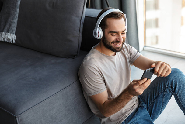 Image of a happy optimistic young man indoors at home listening music with headphones sit on floor chatting by mobile phone. - Φωτογραφία, εικόνα