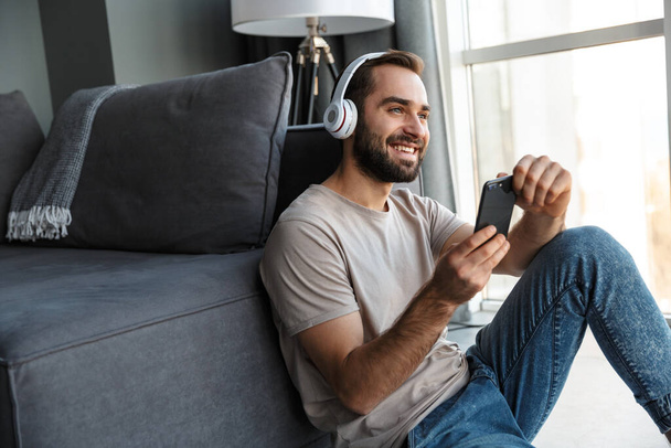 Image of a smiling young man indoors at home listening music with headphones sit on floor chatting by mobile phone. - Фото, зображення