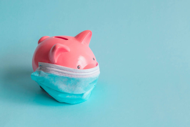 Pink piggy bank, wearing protective facemask, isolated on blue background. Piggy bank with disposable medical mask. Money saving concept during coronavirus pandemic. - Foto, Imagem