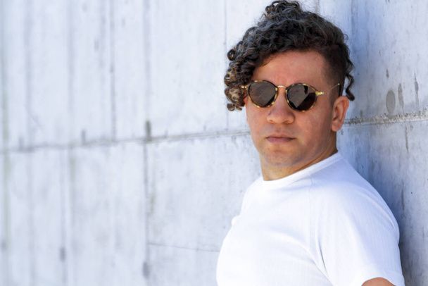 Portrait of a stylish man with curly hair and sunglasses leaning against a concrete wall - Фото, изображение