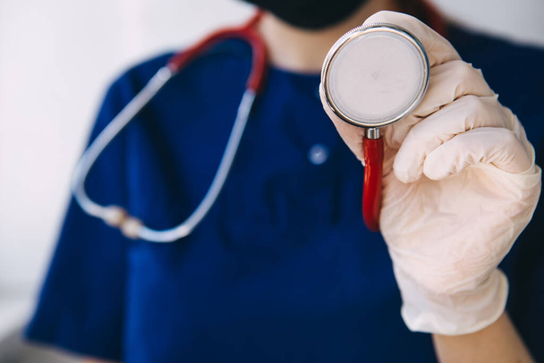 fiatal orvos egy sebészeti öltöny, kesztyű és maszk tart egy piros sztetoszkóp, hallgat egy beteg koronavírus - Fotó, kép