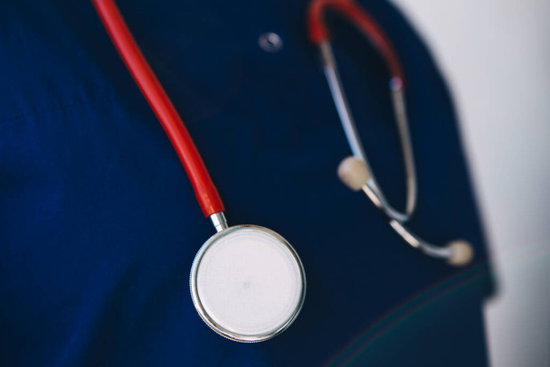 молодой врач в хирургическом костюме, перчатках и маске держит красный стетоскоп, слушает пациента с коронавирусом
 - Фото, изображение
