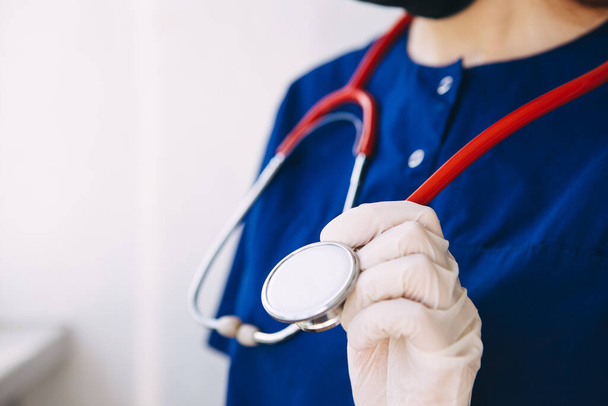 молодой врач в хирургическом костюме, перчатках и маске держит красный стетоскоп, слушает пациента с коронавирусом
 - Фото, изображение