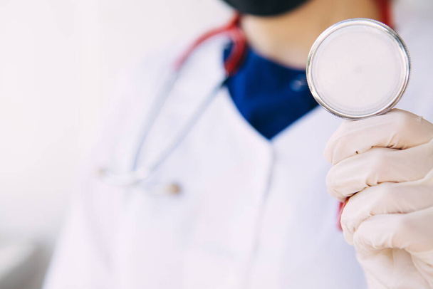 médico joven con un abrigo médico, guantes y máscara sostiene en la mano un estetoscopio rojo, escucha a un paciente con coronavirus
 - Foto, imagen