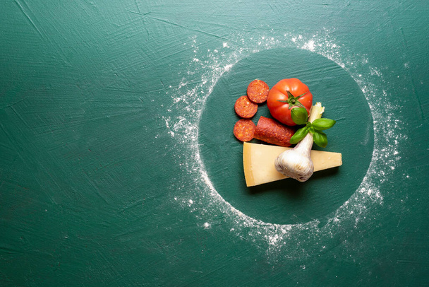 Ingredientes de comida para fazer uma pizza pepperoni em uma mesa verde, acima vista. Ingredientes de pizza dentro de um círculo de farinha
 - Foto, Imagem