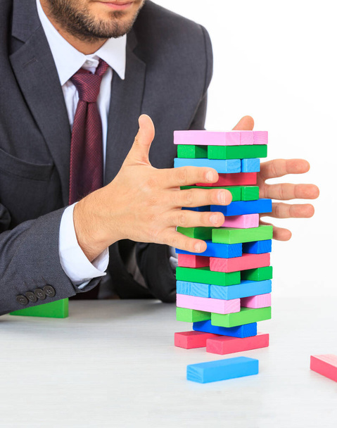 Torre de blocos coloridos em uma mesa de escritório branca. Um homem de fato a tentar manter o equilíbrio. Estratégia de negócios, risco, conceito de construção
 - Foto, Imagem