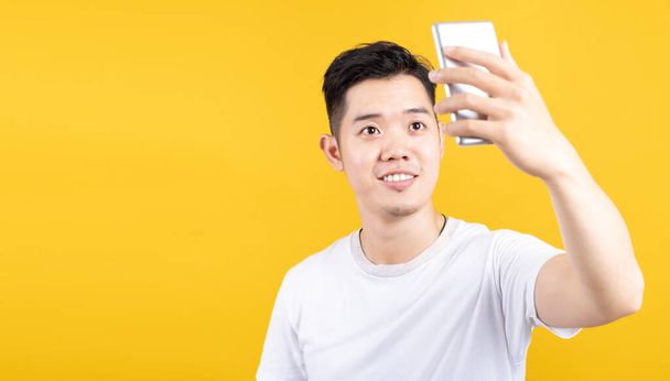 Portrait happy young asian man taking selfie with mobile phone camera smiling with copy space wearing white t-shirt on yellow background isolated studio shot. - Φωτογραφία, εικόνα
