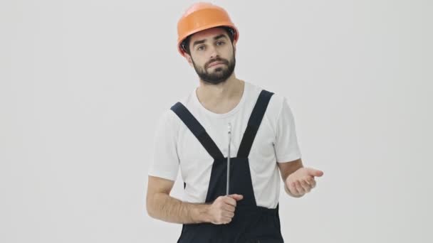 Concentrated young bearded man builder isolated over white wall background in helmet with wrench - Imágenes, Vídeo