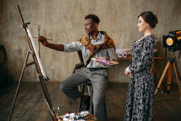 Kobieta patrzy jak malarz z paletą rysuje pędzlem. Mężczyzna siedzący przy sztaludze, wnętrze pracowni sztuki na tle, mistrz kreatywności w warsztacie - Zdjęcie, obraz