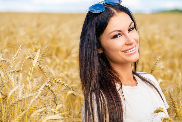Portrait of a young brunette woman on a background of golden wheat field, summer outdoors - Photo, Image