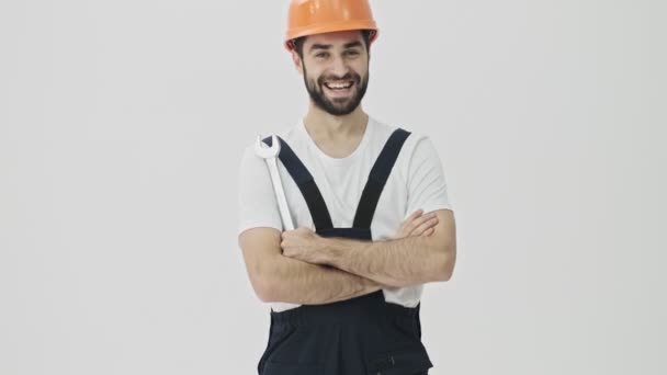 Happy positive young bearded man builder isolated over white wall background in helmet with wrench - 映像、動画