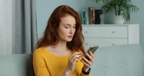 Close up of Caucasian young red-haired woman sitting on sofa in living room and shopping online on smartphone. Pretty female shopper buying on phone and paying with credit card. Self-isolation at home - 映像、動画