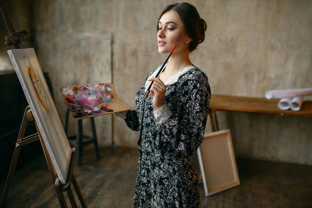 Female painter with brush and palette poses in art studio. Artist at her workplace, creative master at the easel in workshop - Photo, Image