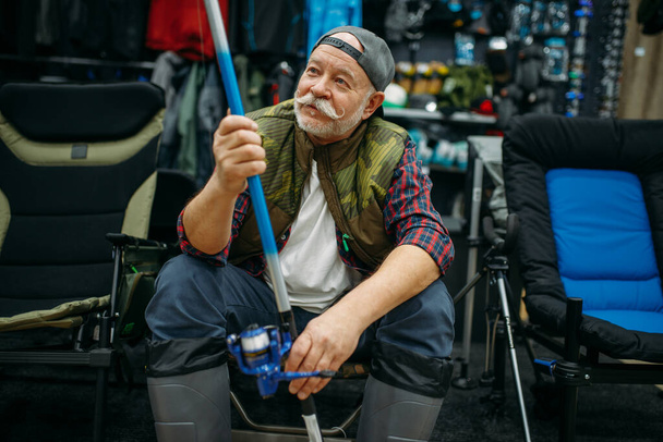 Lastik botlu erkek balıkçı, oltayı balıkçı dükkanında tutuyor, birinci şahıs manzaralı. Balık yakalamak ve avlamak için ekipman ve araçlar, vitrindeki aksesuar seçimi - Fotoğraf, Görsel