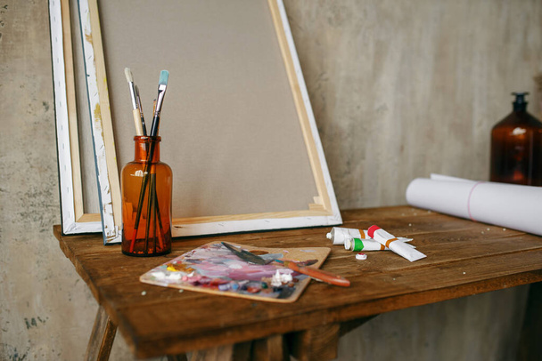 Paints on palette, brush in bottle, canvas, nobody. Painter tools on the table in art studio, equipment on artist workplace, paintbrush, creative atelier or workshop - Fotoğraf, Görsel