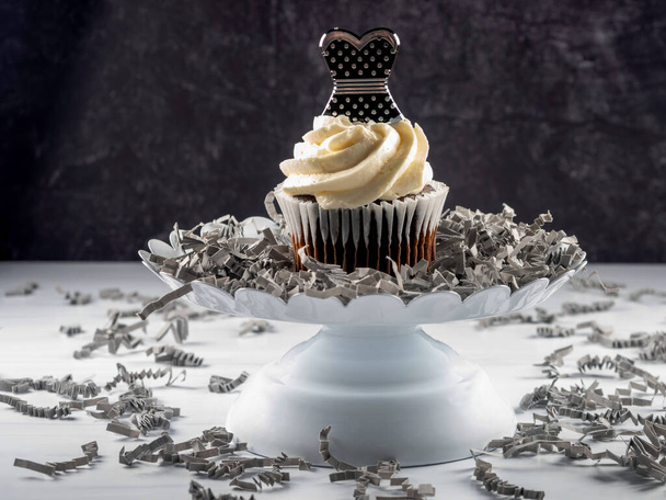 Üstü beyaz kremalı çikolatalı kek ve üstünde siyah ve gümüş benekli bir balo elbisesi, üzerinde gri kağıt parçaları serpiştirilmiş, yükseltilmiş beyaz bir tabakta oturuyor.. - Fotoğraf, Görsel