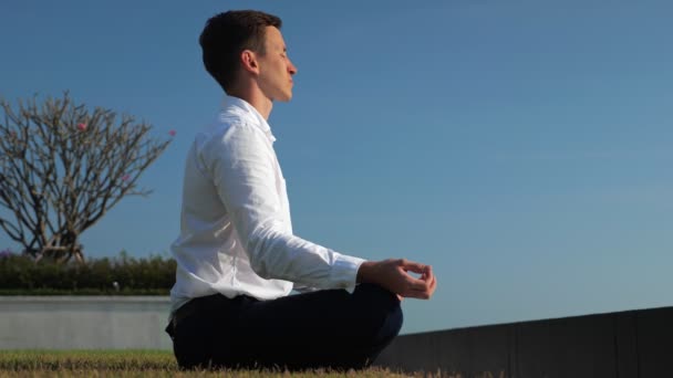 fiatal üzletember fehér ingben meditál napfény alatt szemben zöld domb teraszon - Felvétel, videó