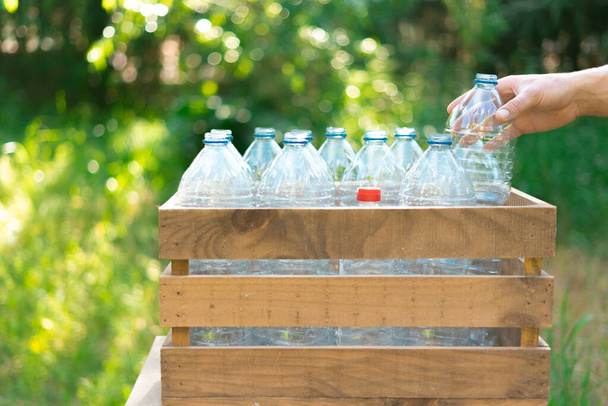 Opětovné použití koncepce recyklace plastových lahví. Mužská ruka zanechává plastovou láhev v recyklované dřevěné krabici s plastovými láhvemi na vodu bez víčka s pěkným západem slunce na zahradě - Fotografie, Obrázek