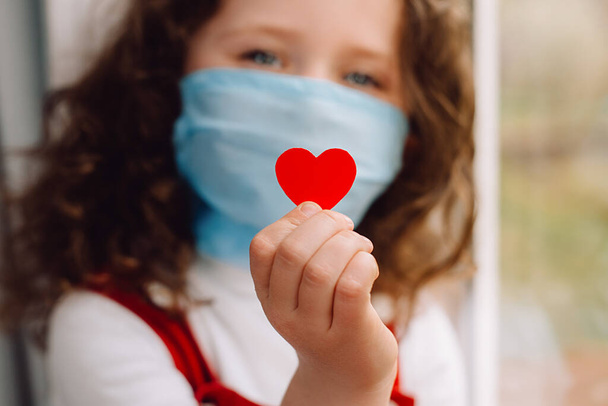 Cute preschool little girl in protective medical masks sitting on sill, holding red heart a way to show appreciation and to thank all essential employees during Covid-19 pandemics. Selective focus - Foto, immagini