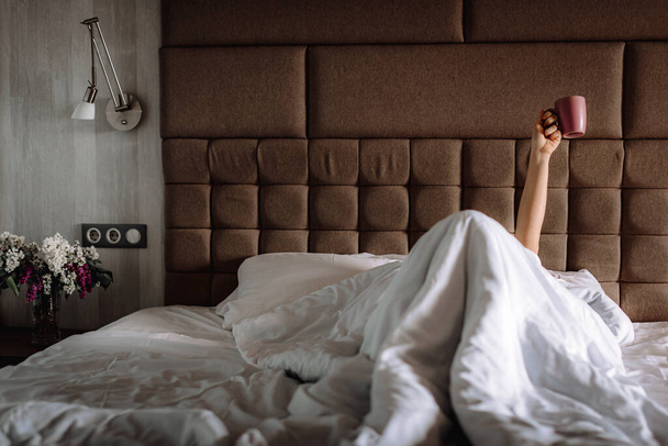 Mujer en la cama, debajo de una manta, con la mano levantada, sosteniendo una taza de café. El ritual matutino de despertar
 - Foto, Imagen