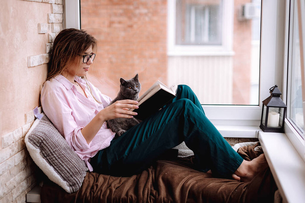 girl in glasses reads a book near the window. Young woman is relaxing on the balcony with her kitten - 写真・画像
