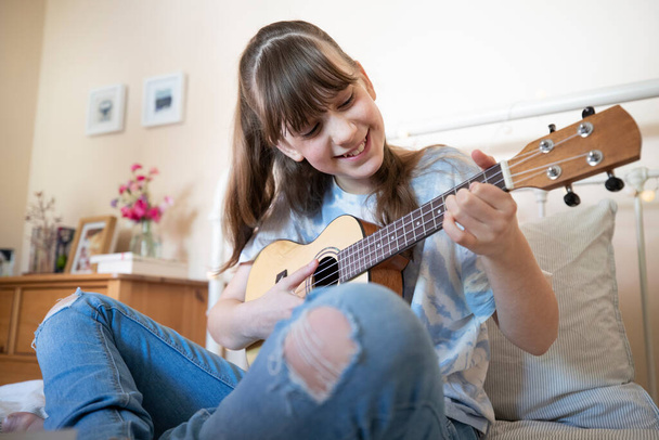 Girl Learning To Play Ukulele Sitting On Bed In Bedroom - Foto, afbeelding