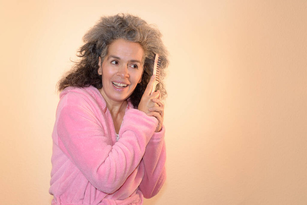 A smiling woman over fifty wearing a pink bathrobe is combing her graying curly hair. Gradient terracotta background. - Photo, Image