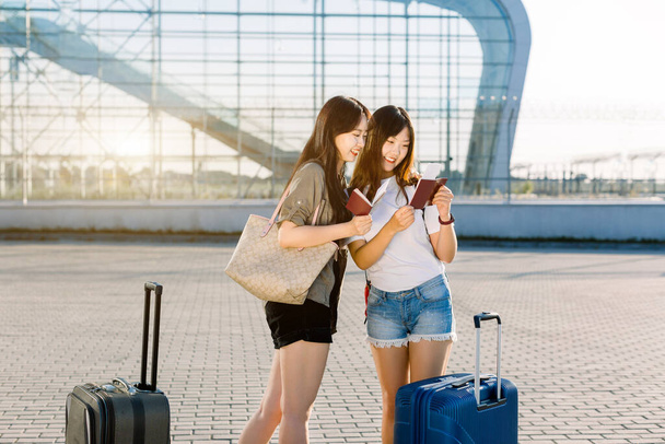 Pasaportlarını ve uçuş biletlerini kontrol ederken, modern havaalanı kapısının yanında büyük bavullarıyla bekleyen, sıradan giysili iki güzel mutlu Asyalı kadın. Ortak gezi, tatil konsepti - Fotoğraf, Görsel