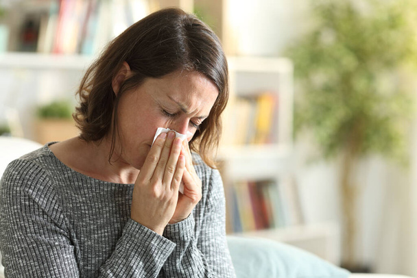 Sick adult woman blowing nose with tissue sitting on the couch at home - Foto, Bild