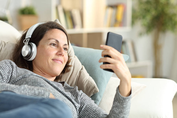 Happy adult lady with headphones listens music on smart phone lying on a couch at home - 写真・画像