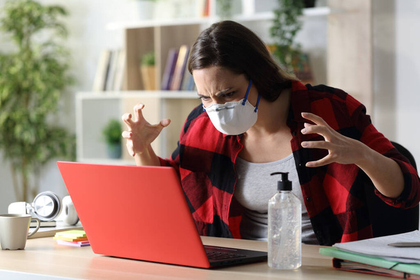 Angry student woman with mask reading news on laptop sitting on a desk on covid-19 confinement at home - 写真・画像