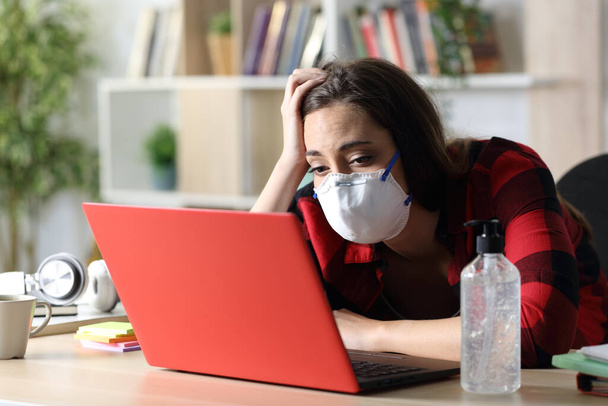 Bored student woman with mask looking at laptop sitting on a desk in coronavirus confinement at home  - Φωτογραφία, εικόνα