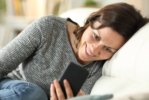 Happy adult woman checking smart phone relaxing sitting on a couch at home - Foto, Imagem