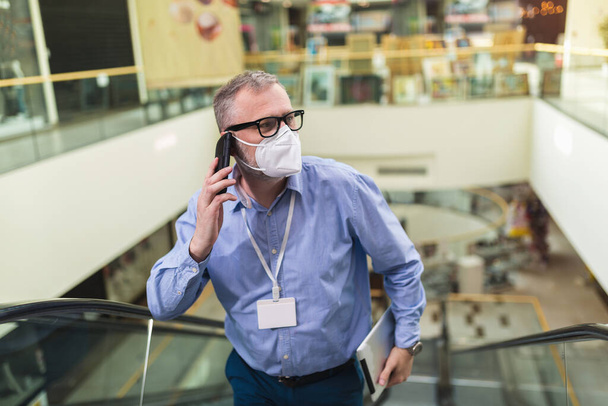 Όμορφος επιχειρηματίας με προστατευτική μάσκα προσώπου και χρήση τηλεφώνου σε κυλιόμενες σκάλες. - Φωτογραφία, εικόνα