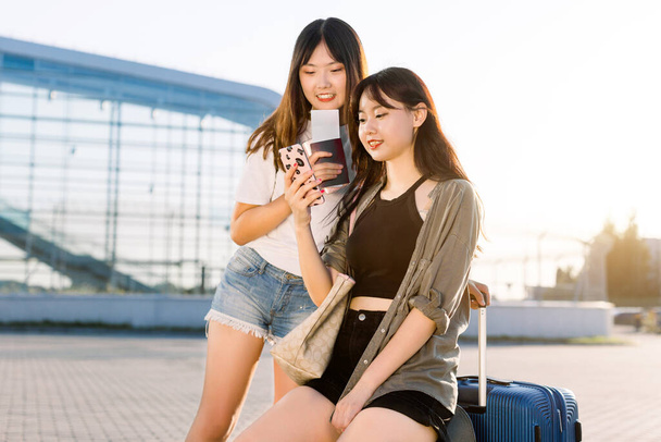 Two excited pretty young asian brunette girls in casual youth clothes, using smartphone to check their flight or enjoying social networks, waiting for their trip on the airport background - Fotoğraf, Görsel