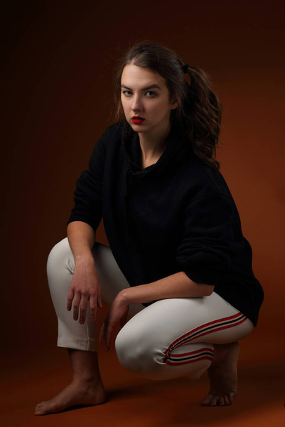 Estúdio retrato de uma menina adolescente bonita em um capuz preto e calças de treino de cor clara
 - Foto, Imagem